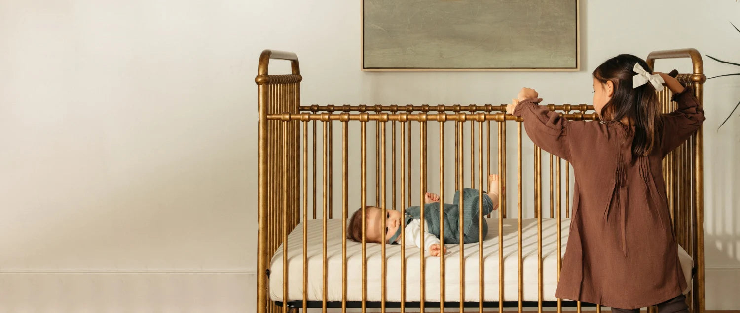 A baby plays in a gold colored iron crib while his sister looks in at him.