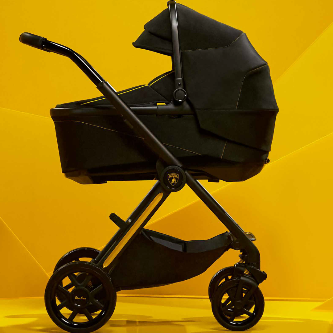 A black baby stroller with a bassinet attachment, shown in profile against a bright yellow geometric studio background.
