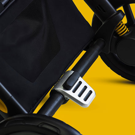 Close-up of a black stroller frame with a silver metal footrest attachment, set against a solid yellow background.