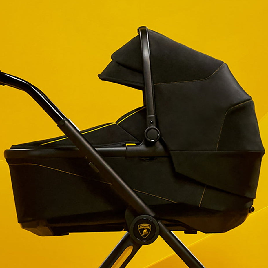 A black baby stroller and bassinet combo with a gold accent ring, shown in profile against a bright yellow geometric studio background.